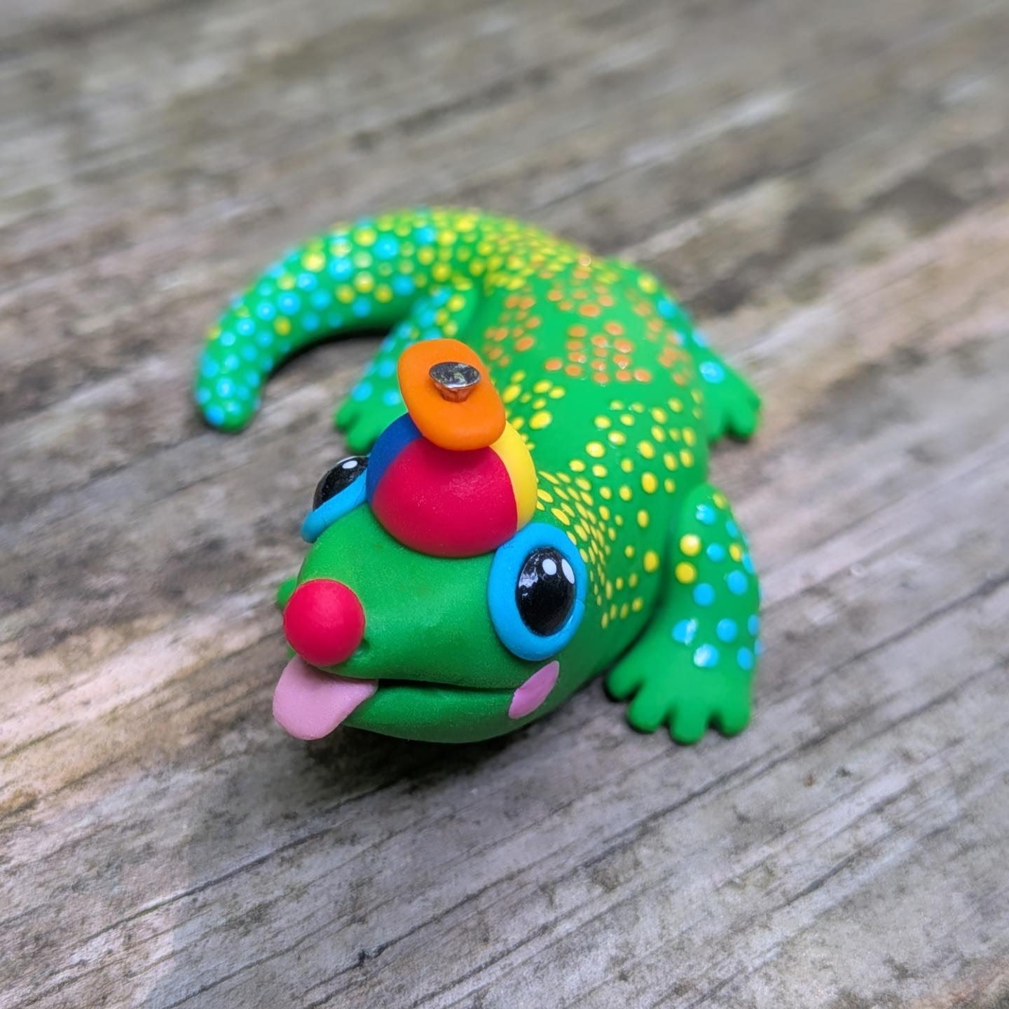 A palm-sized sculpture of a clown-themed gold dust day gecko. It's body is bright green with colorful dots of yellow, green, and blue. It wears a red, yellow, and blue hat with an orange propeller. The lizard has a red clown nose, pink painted cheeks, wide eyes, and it's tongue hangs out in a silly way.