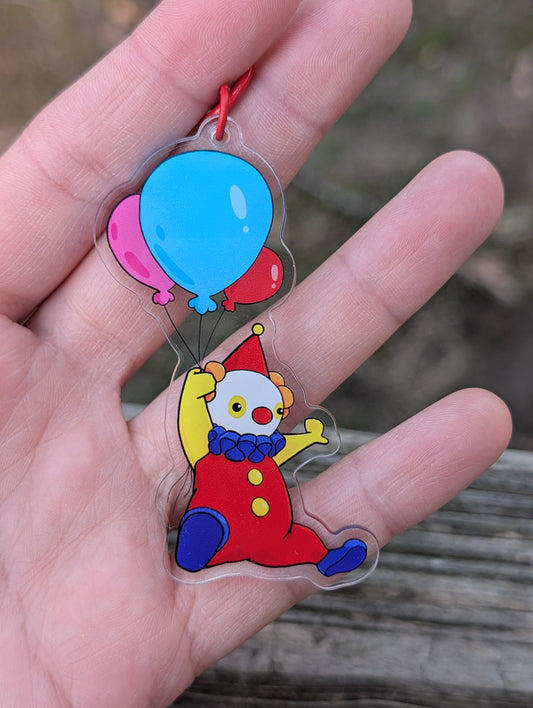 An acrylic keychain of a clown flying with a handful of 3 balloons. The clown has a red suit, yellow arms, blue shoes and frilly collar, and a red hat. The balloons are pink, blue, and red, respectively. The keychain has a red lobster clasp.