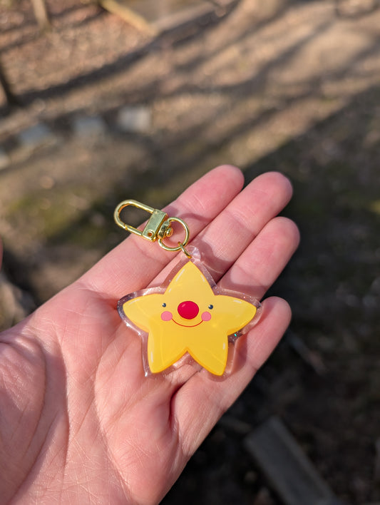 A yellow star-shaped acrylic keychain with a cute red clown nose, pink cheeks, little eyes, and a smiley face, held in a person's hand. The keychain has a gold-tone lobster clasp and a glitter layer.