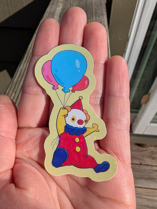 A glossy vinyl sticker of a cartoon clown flying with a handful of 3 balloons. The clown has a red outfit, yellow arms, blue shoes and frilly collar, and a red hat. The balloons are pink, blue, and red. The sticker follows the outline of the clown art, but has a small border. The border/background is pale yellow. The sticker has a holographic glitter effect.