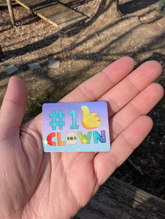 A small, rectangular sticker with rounded corners that reads "#1 CLOWN". The "#1" is teal, the "CLOWN" is rainbow, and there is a big, yellow thumbs-up. The letter O in "CLOWN" is a clown's face. The background is purple and light blue ombre. The sticker is glossy with a holographic star coating.