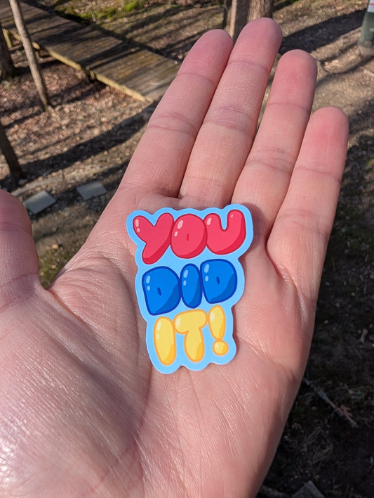 A vinyl sticker that says "YOU DID IT" in balloon lettering. Each letter is stacked on top of each other. The "YOU" is red, "DID" is blue, and "IT" is yellow. The sticker follows the shape of the lettering with a small, blue border around the letters.
