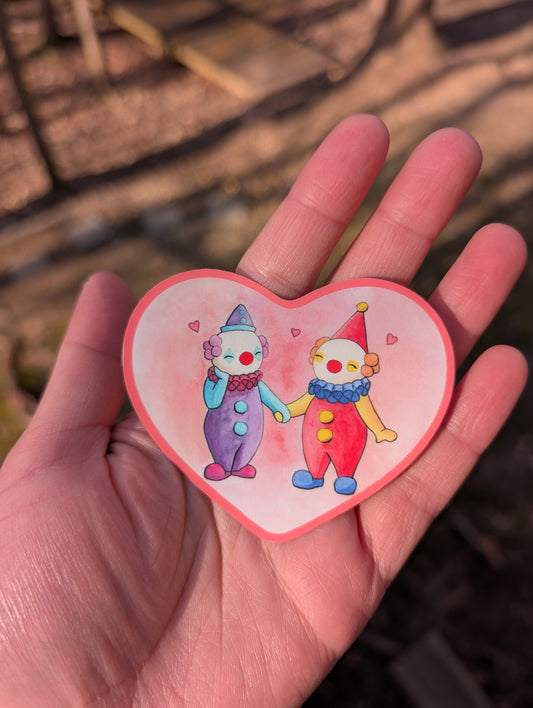 A pink, heart-shaped sticker of two clowns holding hands. One clown has a red, yellow, and blue outfit with orange hair, and the other clown has a purple and blue outfit with purple hair. There are hearts in between them and they look happy. The image was originally hand drawn and water colored, so it has a handmade look to it.