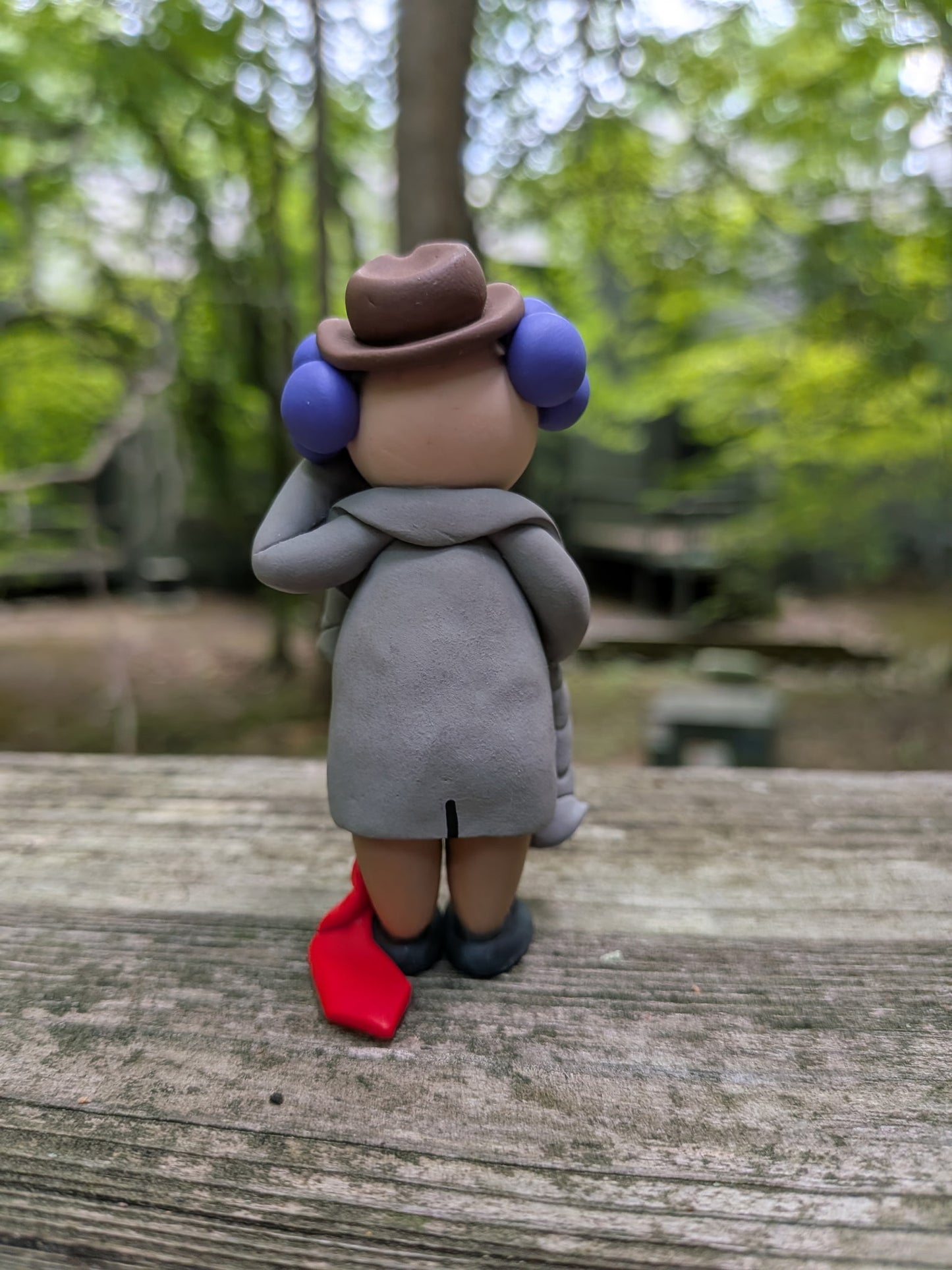 A view of the funny business clown from behind, wearing a brown fedora, oversized grey suit, brown pants, and black shoes. It shows his hair that is bald down the middle. His tie is very long and drags on the ground behind him.