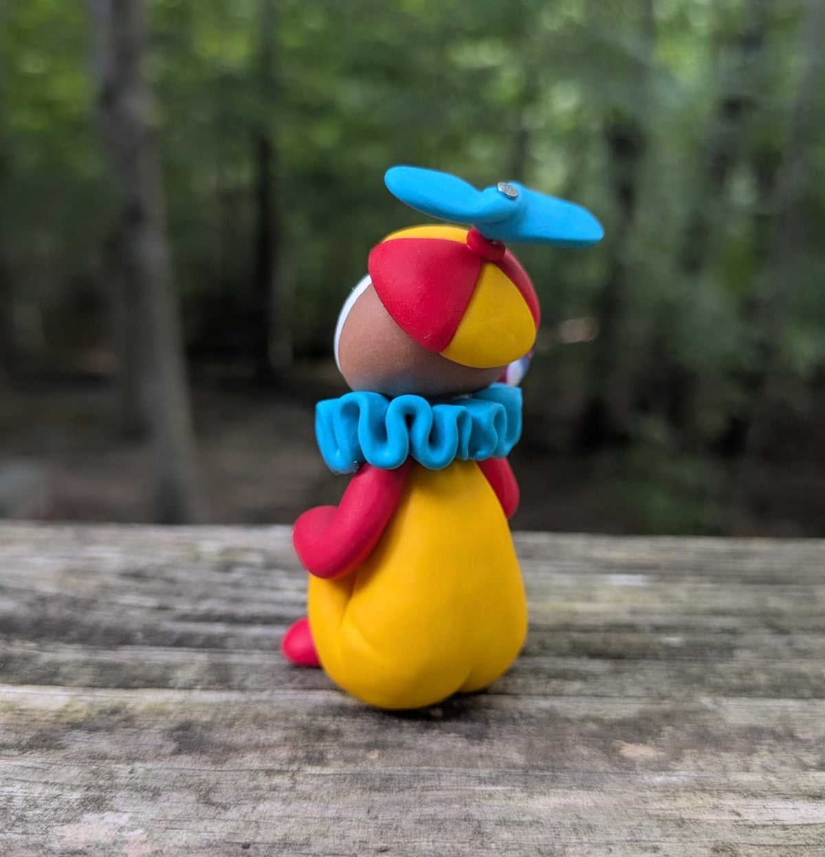 A colorful clay figurine of a clown sitting with a lolipop. The clown has a yellow body, red arms and shoes, a blue ruffled collar and hat, and a white face with red and blue details.