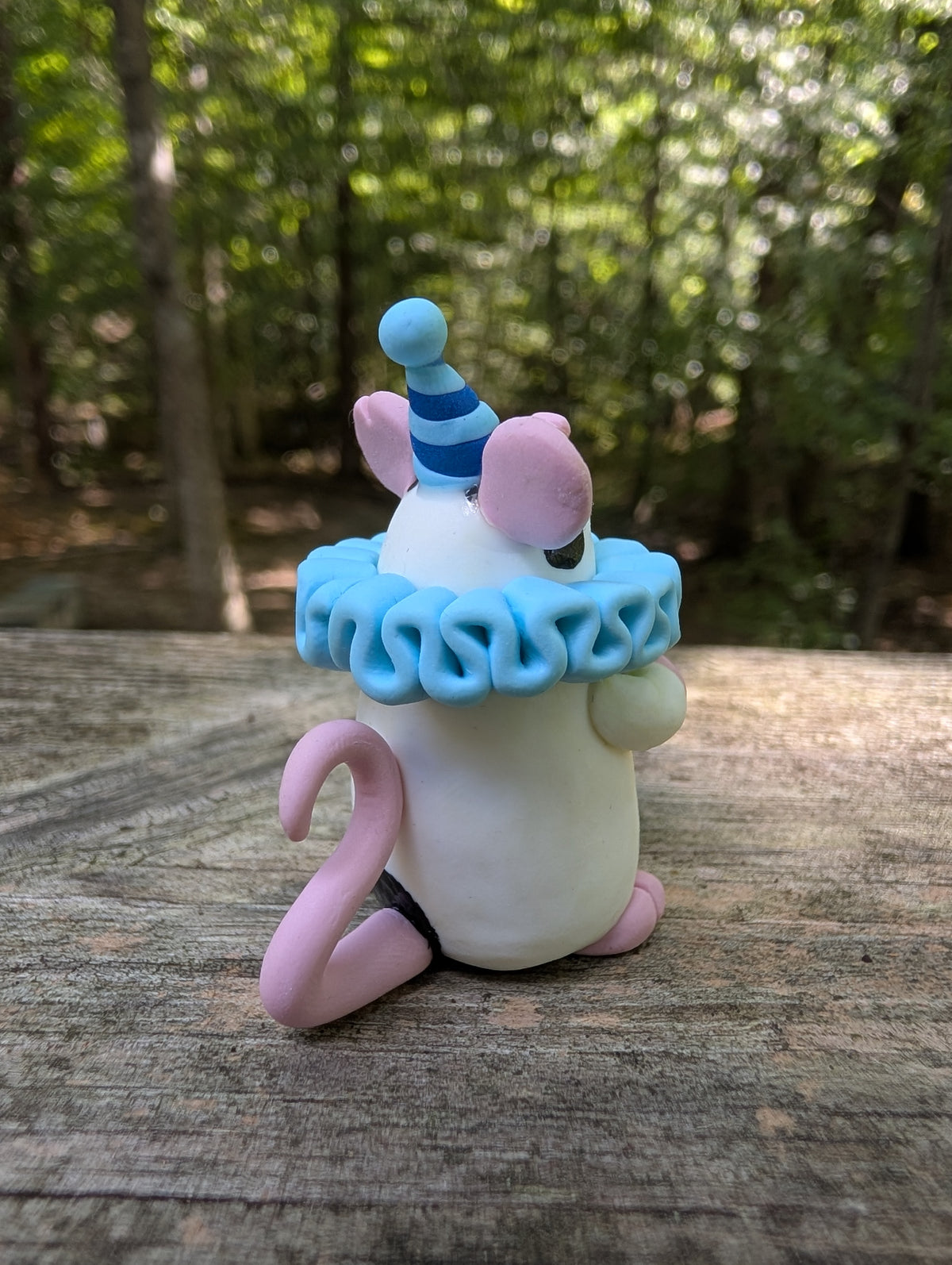 A small white mouse sculpture with pink ears and feet, black markings on the face and butt, wearing a blue striped party hat and a light blue ruffled collar. The mouse stands on it's back legs with it's hands in the air.