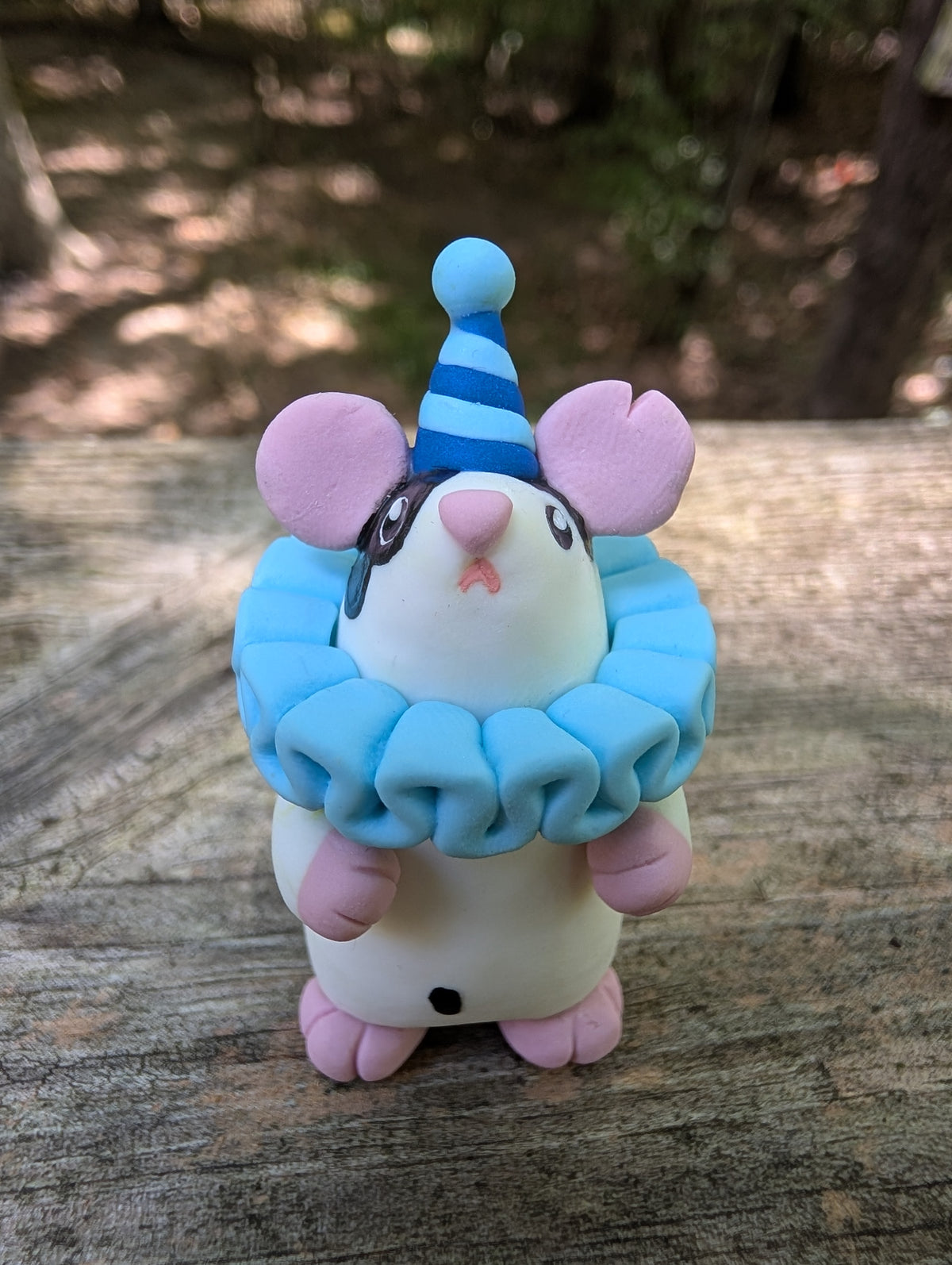 A small white mouse sculpture with pink ears and feet, black markings on the face and belly, wearing a blue striped party hat and a light blue ruffled collar. The mouse stands on it's back legs with it's hands in the air.