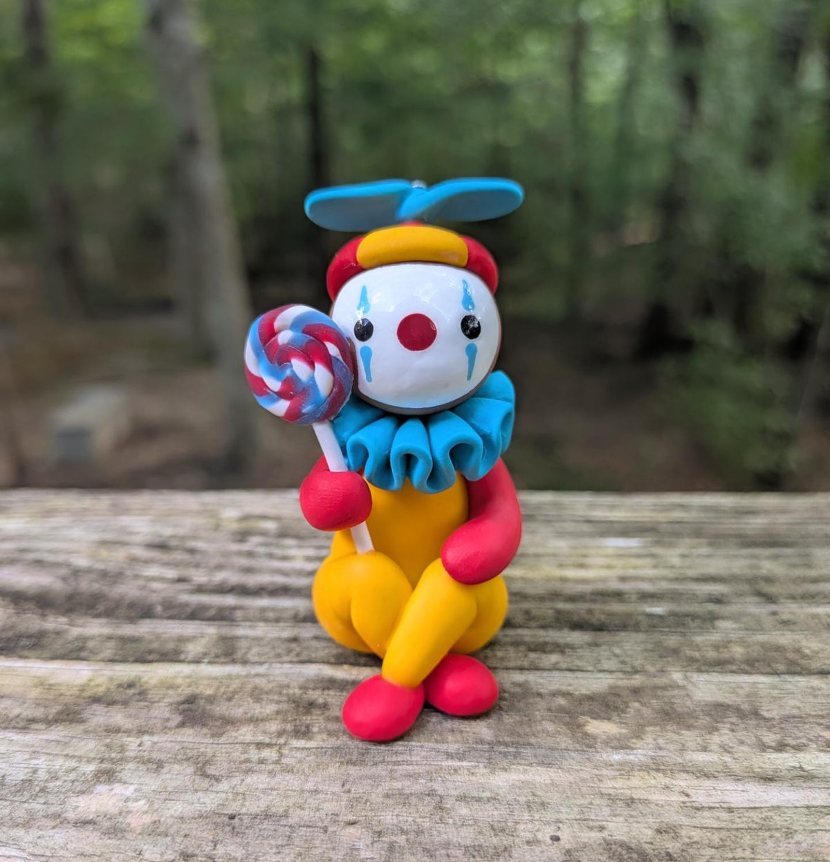 A clown sculpture. The clown yellow, red, and blue. They are holding a large swirly lolipop and sitting on the floor with their legs crossed.