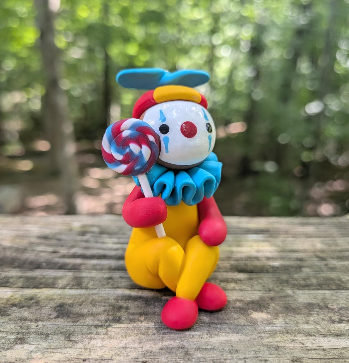 A colorful clay figurine of a clown sitting with a lolipop. The clown has a yellow body, red arms and shoes, a blue ruffled collar and hat, and a white face with red and blue details.