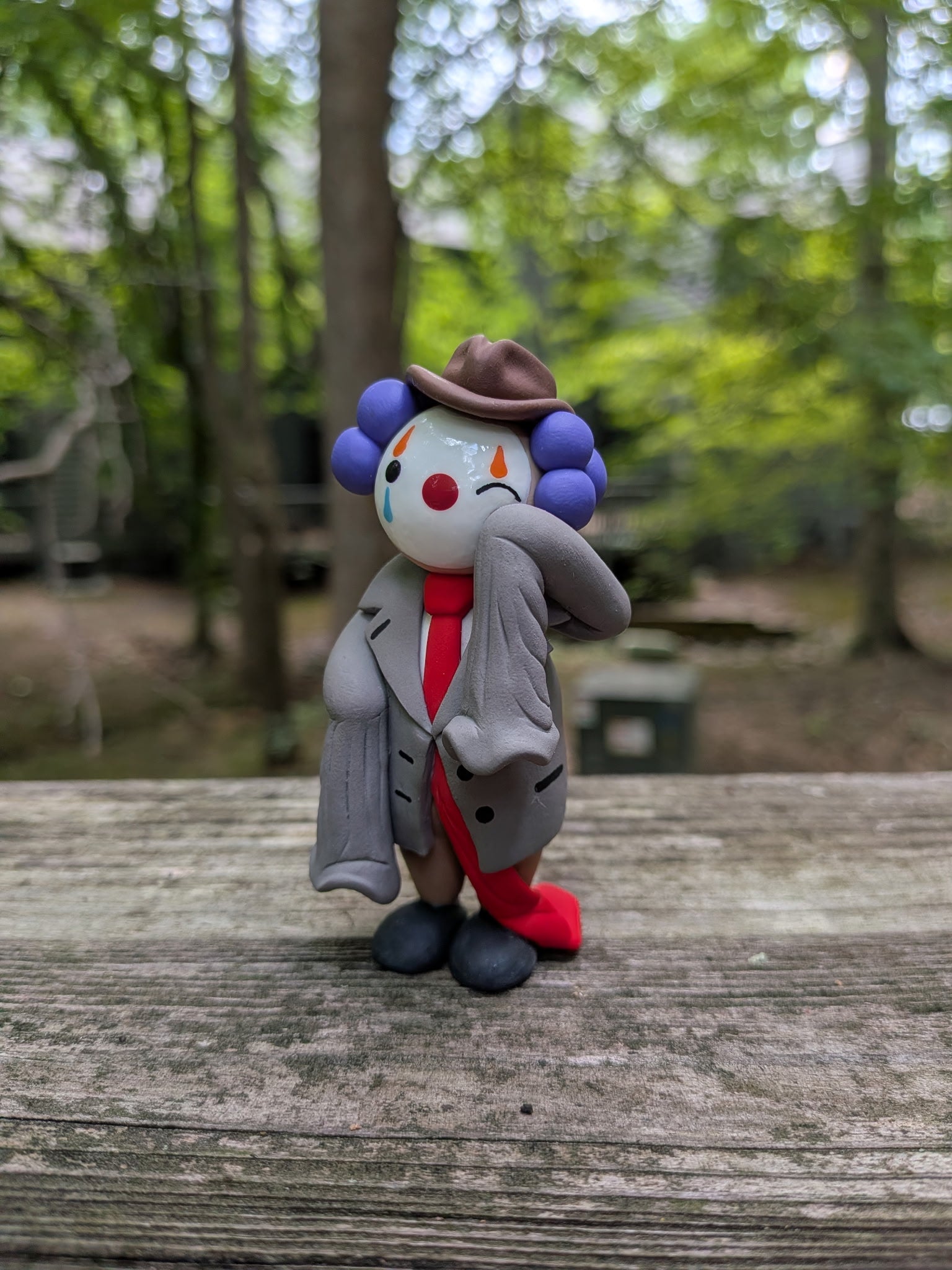 A palm sized polymer clay figurine of a clown dressed in oversized business attire. He is wearing a comedically oversized grey suit with sleeves that dangle as he wipes a tear from his left eye. His long red tie drags on the ground behind him. He is wearing big black shoes, brown pants,and a brown pinch front fedora. He has purple hair made of three large clay balls on either side of his head. He has white face paint, a red nose, and orange and blue eye paint. He has tear drops on the underside of his eyes.