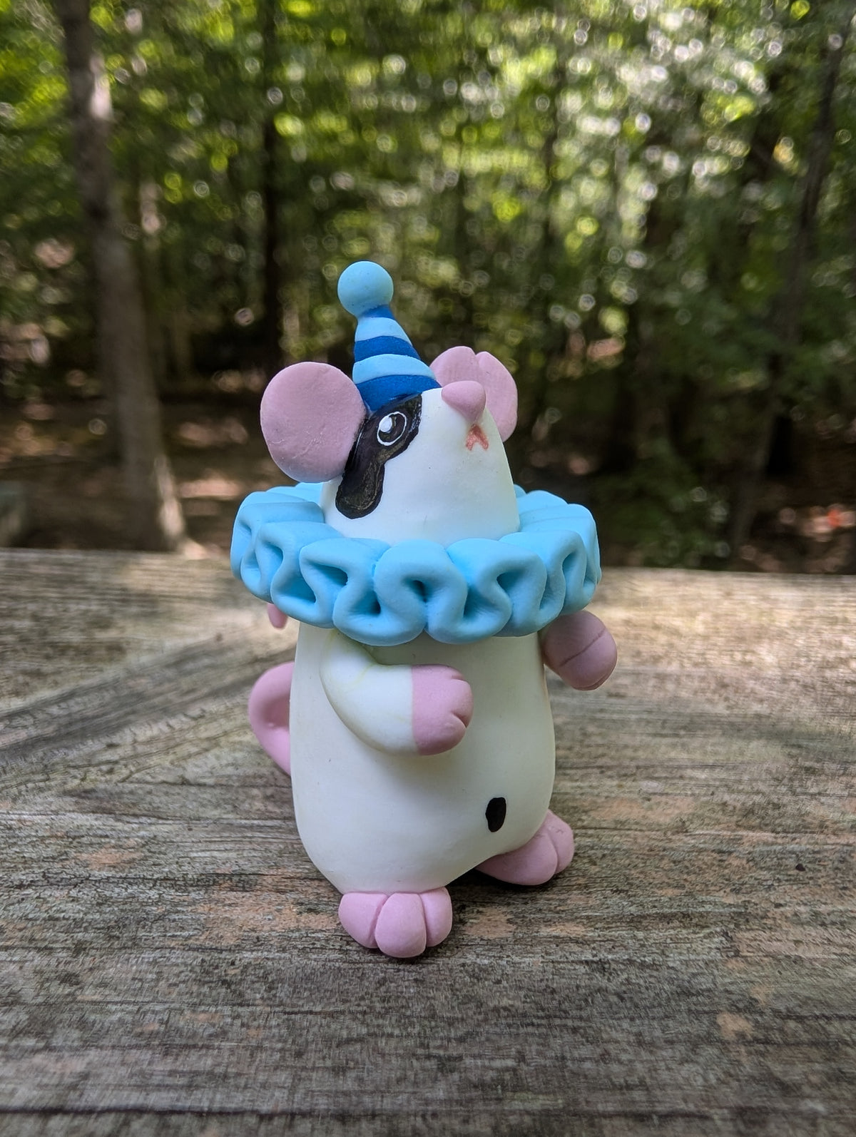 A small white mouse sculpture with pink ears and feet, black markings on the face and belly, wearing a blue striped party hat and a light blue ruffled collar. The mouse stands on it's back legs with it's hands in the air.