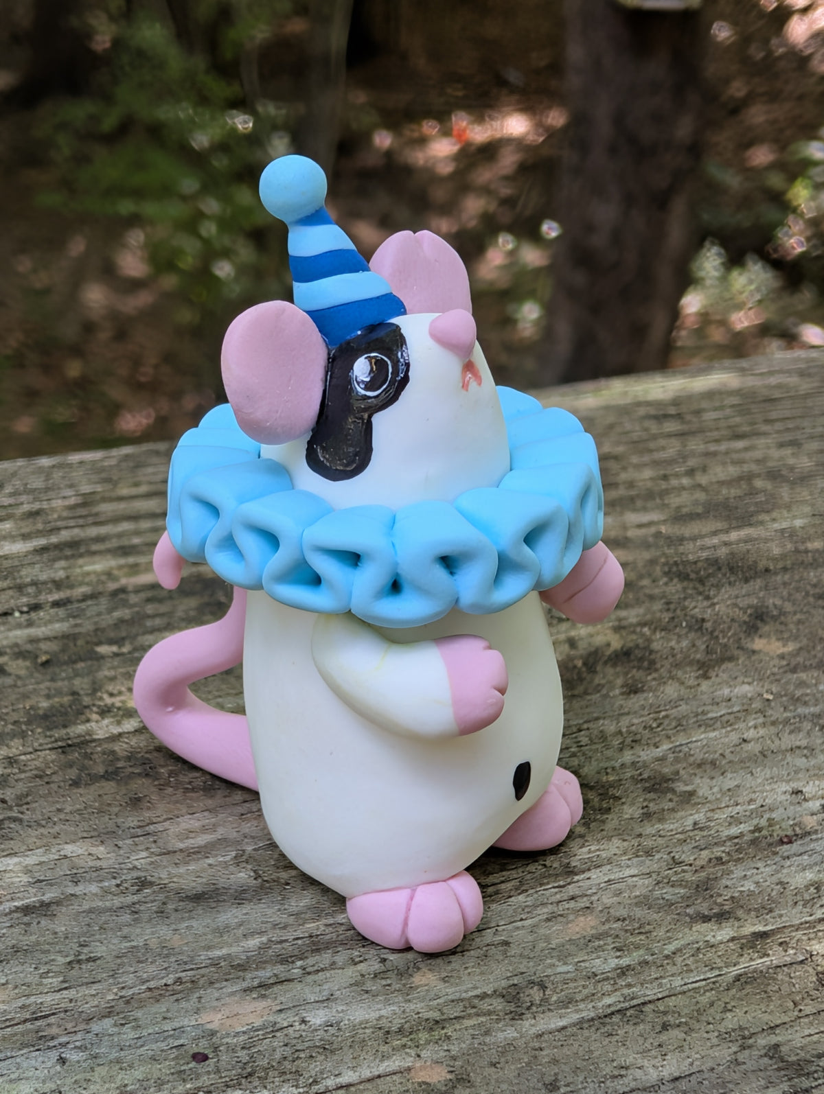 A small white mouse sculpture with pink ears and feet, black markings on the face and belly, wearing a blue striped party hat and a light blue ruffled collar. The mouse stands on it's back legs with it's hands in the air.