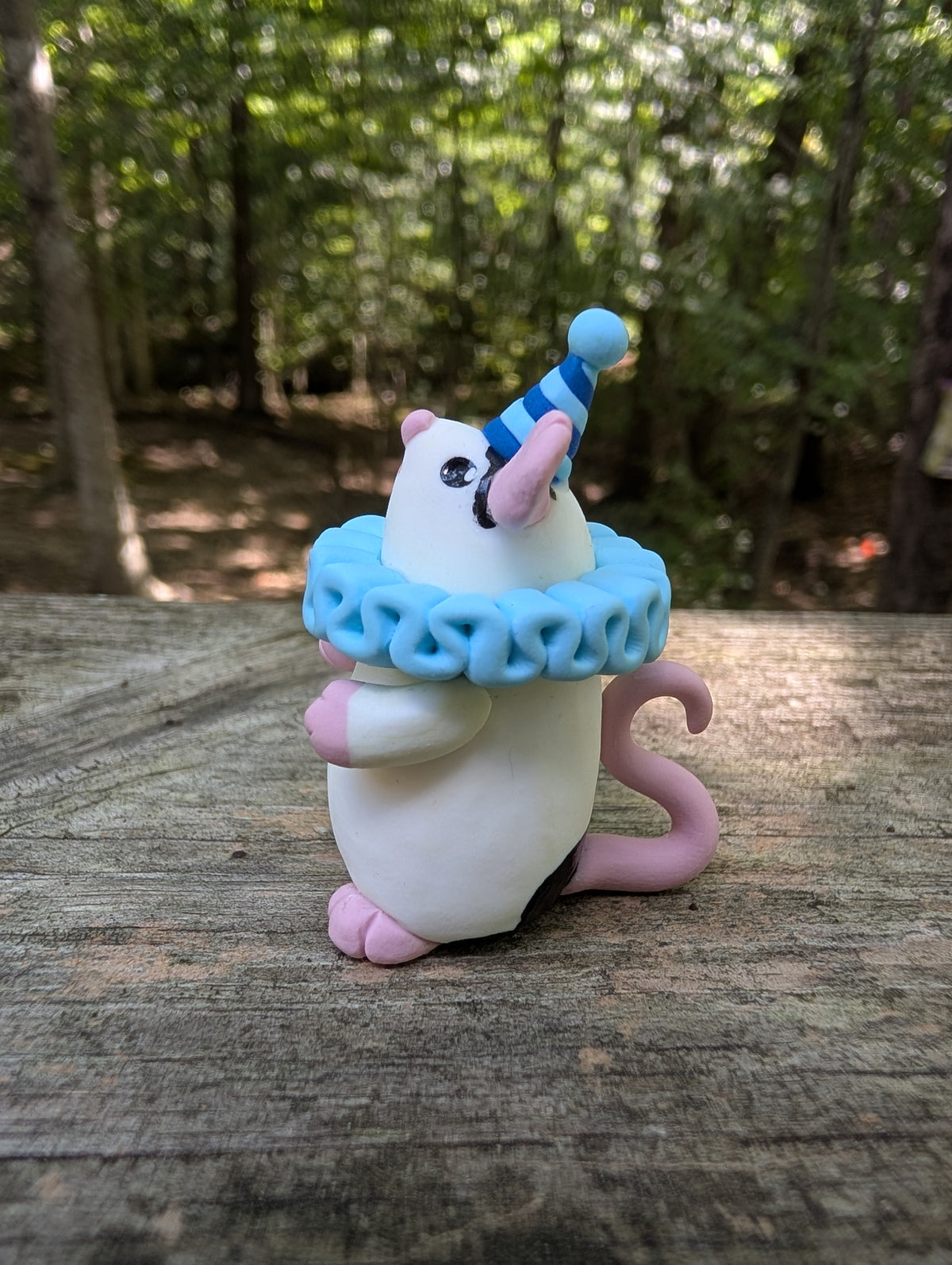 A small white mouse sculpture with pink ears and feet, black markings on the face and belly, wearing a blue striped party hat and a light blue ruffled collar. The mouse stands on it's back legs with it's hands in the air.