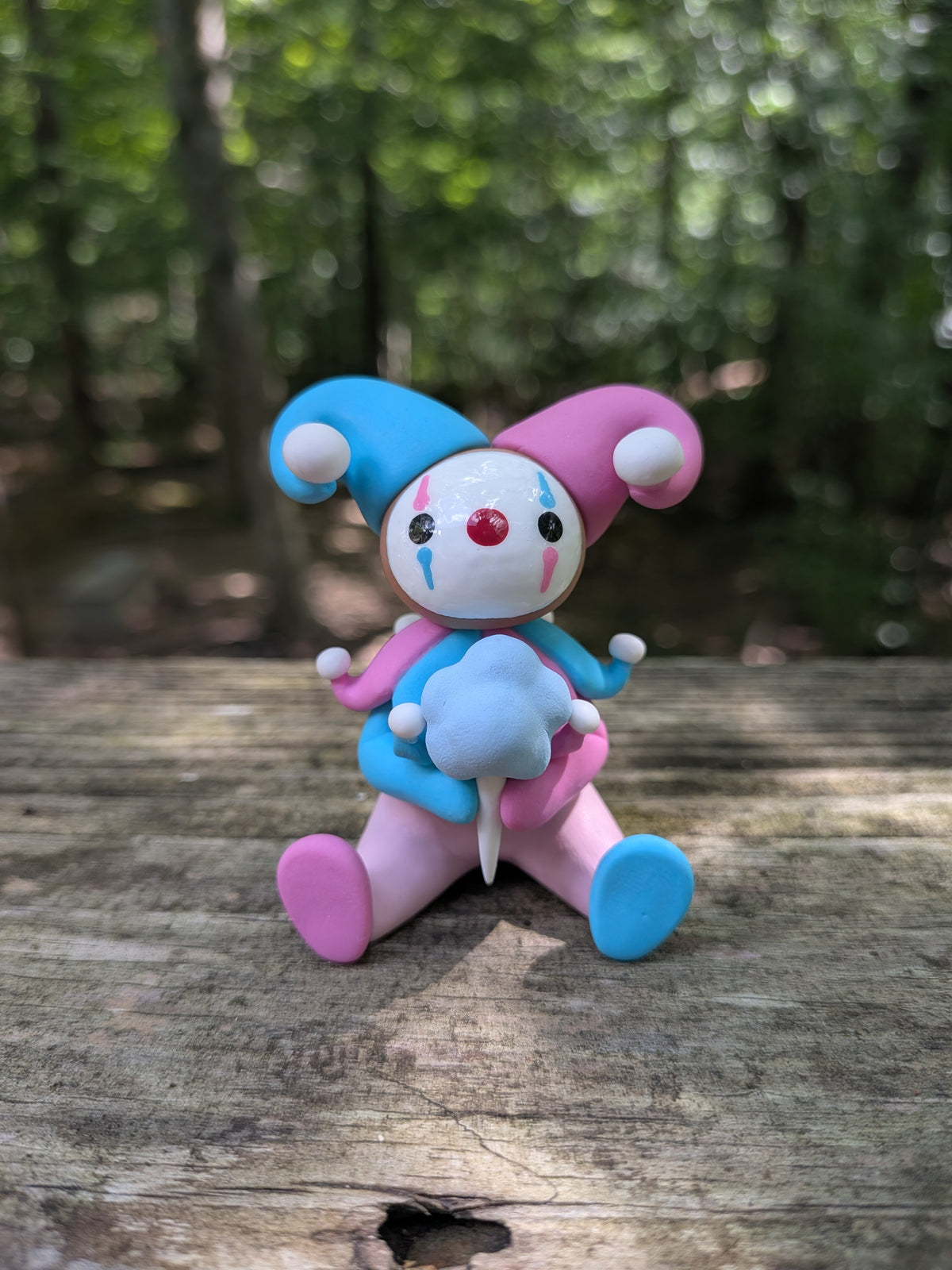 A sculpture of a pink and blue jester clown sitting with a blue cotton candy. He has alternating pink and blue elements and a big jester hat.