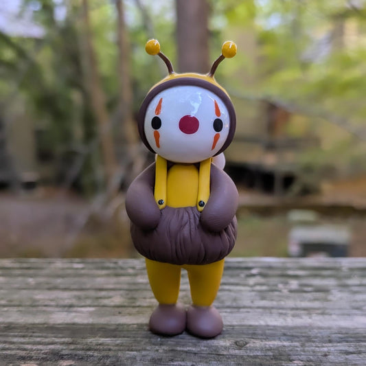 A sculpture of a clown dressed as a bee. He is wearing a yellow jumpsuit with a puffy brown stripe on his lower torso. He has brown shoes and arms, suspenders, and a tight fitting yellow cap with bee antenna.