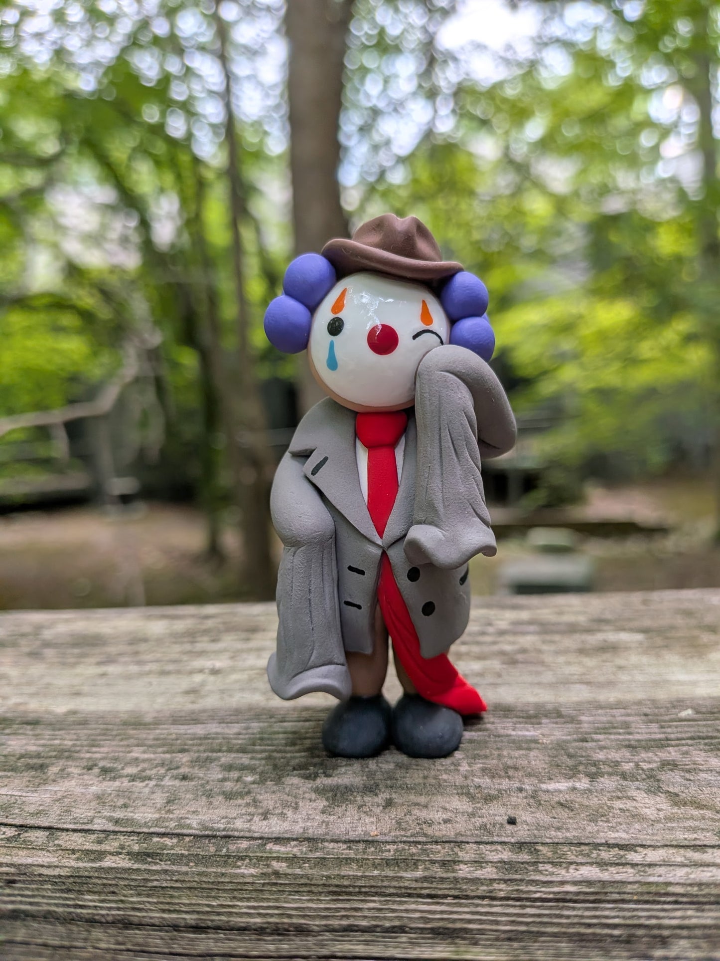 A palm sized polymer clay figurine of a clown dressed in oversized business attire. He is wearing a comedically oversized grey suit with sleeves that dangle as he wipes a tear from his left eye. His long red tie drags on the ground behind him. He is wearing big black shoes, brown pants,and a brown pinch front fedora. He has purple hair made of three large clay balls on either side of his head. He has white face paint, a red nose, and orange and blue eye paint. He has tear drops on the underside of his eyes.