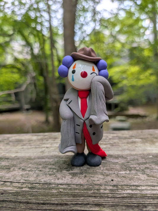 A palm sized polymer clay figurine of a clown dressed in oversized business attire. He is wearing a comedically oversized grey suit with sleeves that dangle as he wipes a tear from his left eye. His long red tie drags on the ground behind him. He is wearing big black shoes, brown pants,and a brown pinch front fedora. He has purple hair made of three large clay balls on either side of his head. He has white face paint, a red nose, and orange and blue eye paint. He has tear drops on the underside of his eyes.
