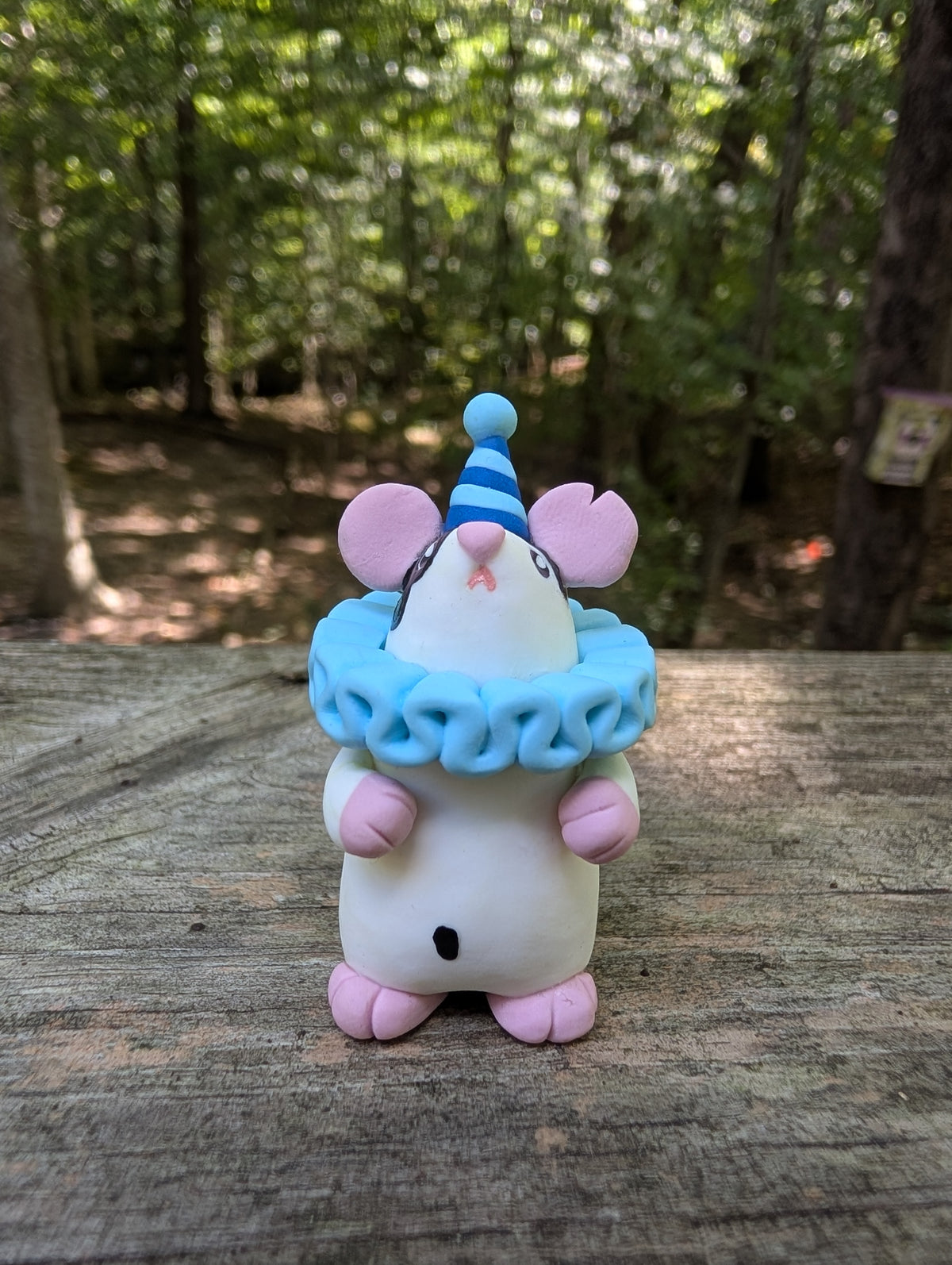 A small white mouse sculpture with pink ears and feet, black markings on the face and belly, wearing a blue striped party hat and a light blue ruffled collar. The mouse stands on it's back legs with it's hands in the air.
