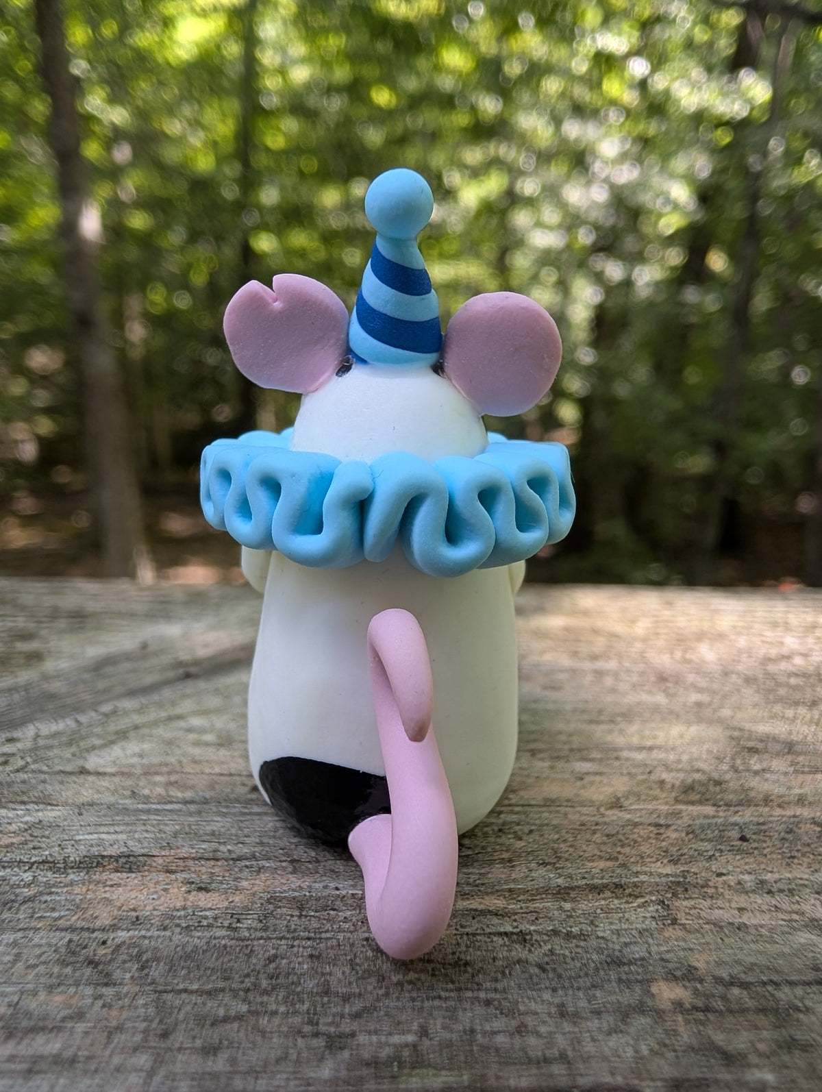 A small white mouse sculpture with pink ears, feet, and tail, black markings on the head and butt, wearing a blue striped party hat and a light blue ruffled collar. This is an angle from the back.