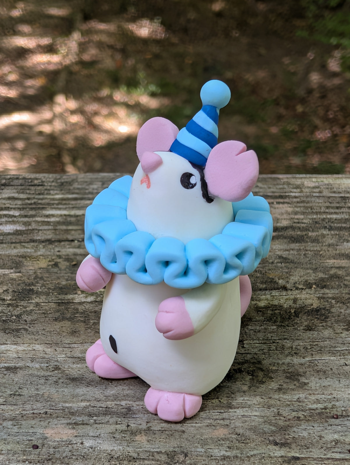 A small white mouse sculpture with pink ears and feet, black markings on the face and belly, wearing a blue striped party hat and a light blue ruffled collar. The mouse stands on it's back legs with it's hands in the air.