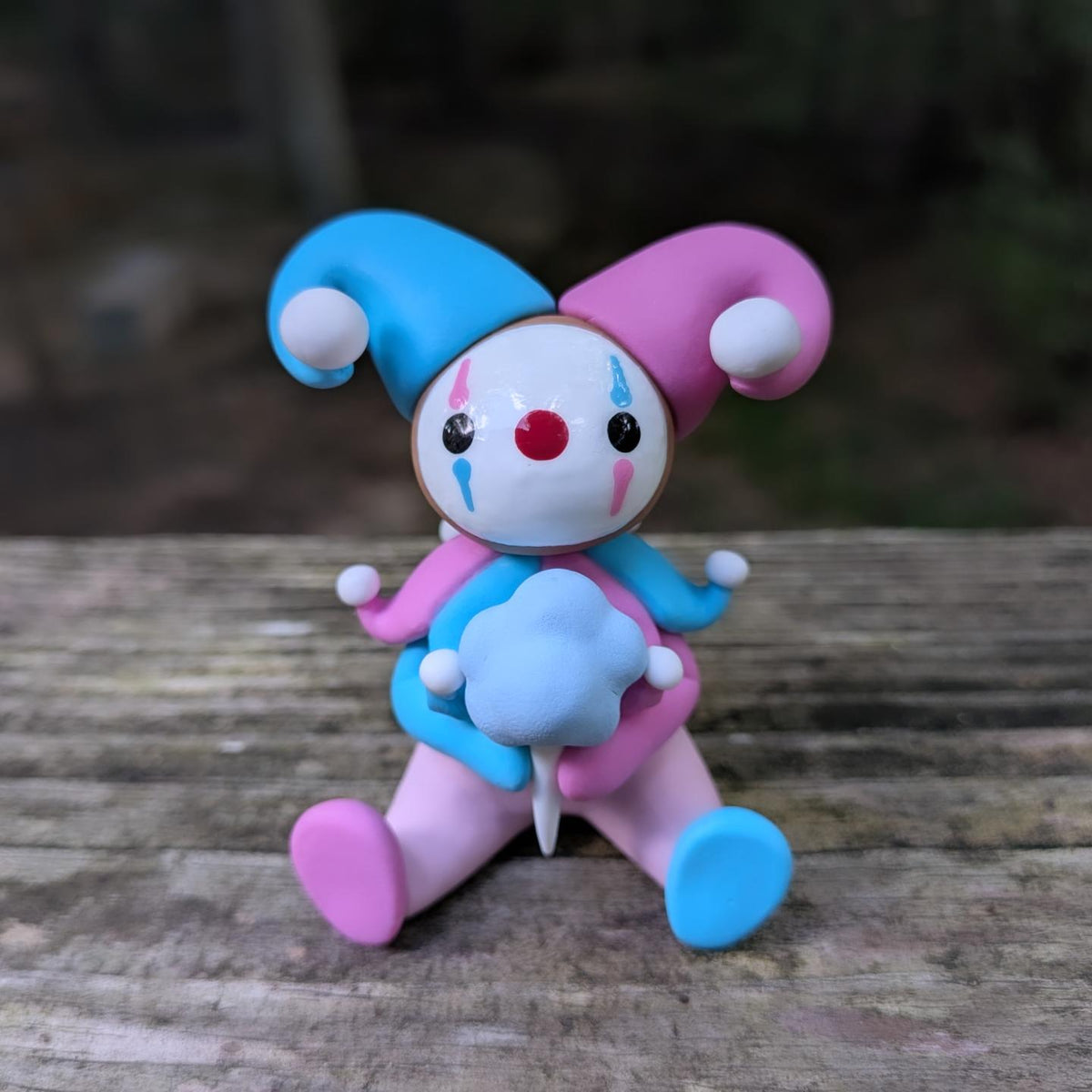 A sculpture of a pink and blue jester clown sitting with a blue cotton candy. He has alternating pink and blue elements and a big jester hat.