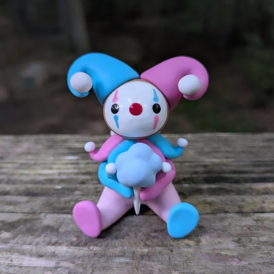 A sculpture of a pink and blue jester clown sitting with a blue cotton candy. He has alternating pink and blue elements and a big jester hat.