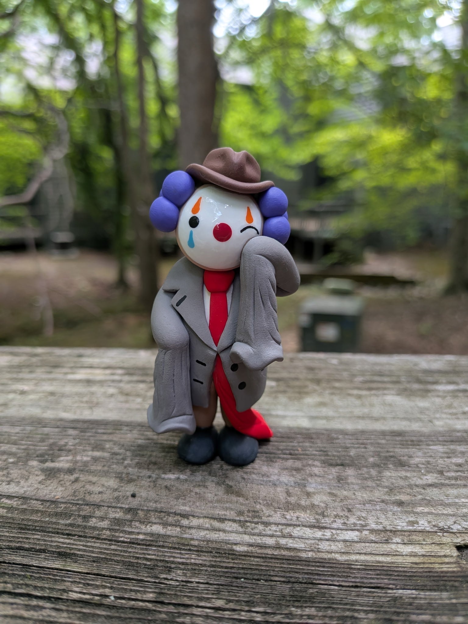 A palm sized polymer clay figurine of a clown dressed in oversized business attire. He is wearing a comedically oversized grey suit with sleeves that dangle as he wipes a tear from his left eye. His long red tie drags on the ground behind him. He is wearing big black shoes, brown pants,and a brown pinch front fedora. He has purple hair made of three large clay balls on either side of his head. He has white face paint, a red nose, and orange and blue eye paint. He has tear drops on the underside of his eyes.