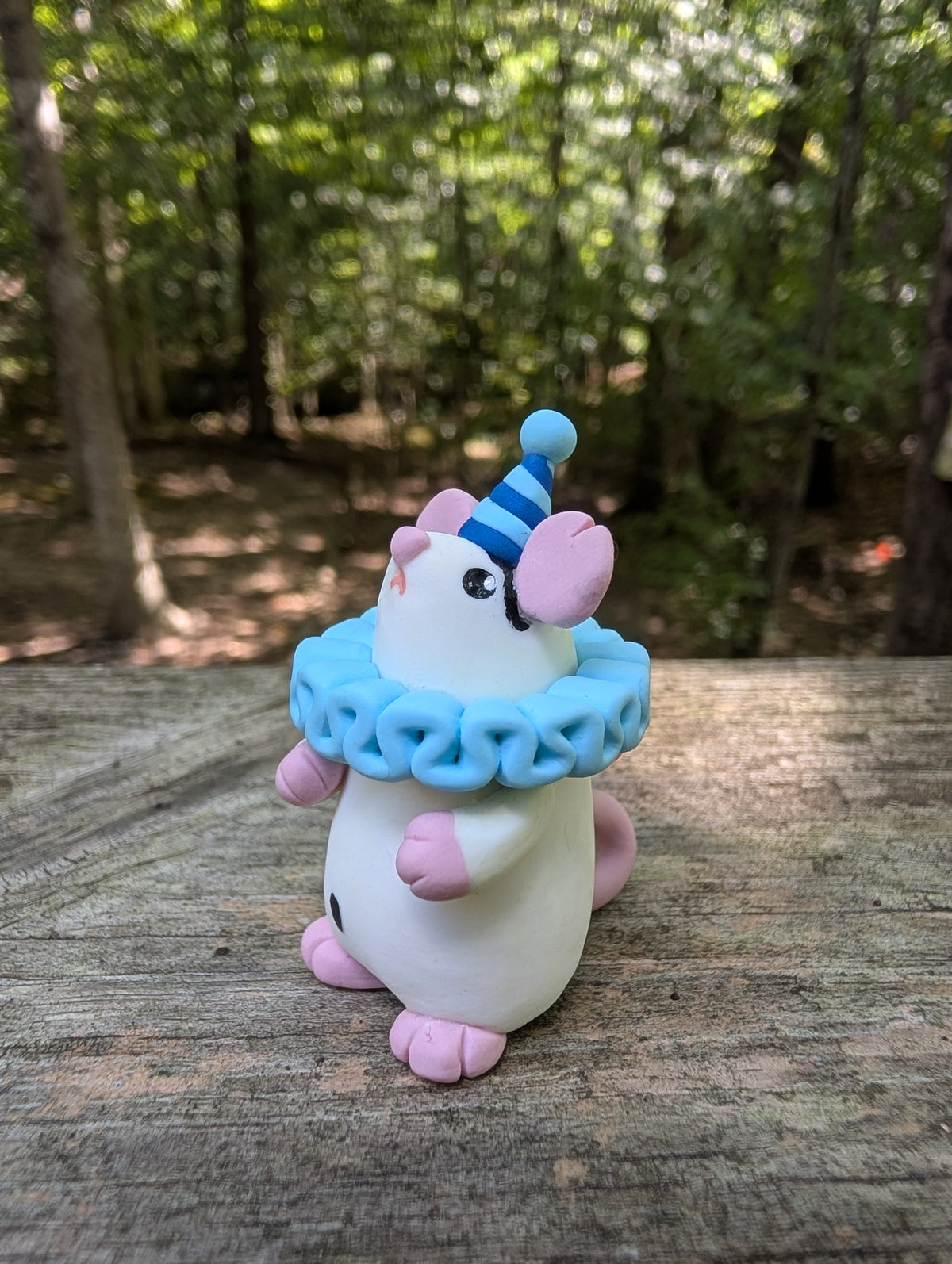 A small white mouse sculpture with pink ears and feet, black markings on the face and belly, wearing a blue striped party hat and a light blue ruffled collar. The mouse stands on it's back legs with it's hands in the air.