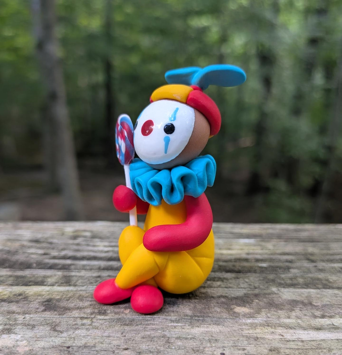 A colorful clay figurine of a clown sitting with a lolipop. The clown has a yellow body, red arms and shoes, a blue ruffled collar and hat, and a white face with red and blue details.