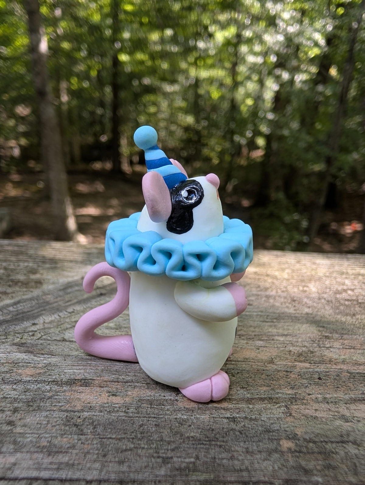 A small white mouse sculpture with pink ears and feet, black markings on the face, wearing a blue striped party hat and a light blue ruffled collar. The mouse stands on it's back legs with it's hands in the air.