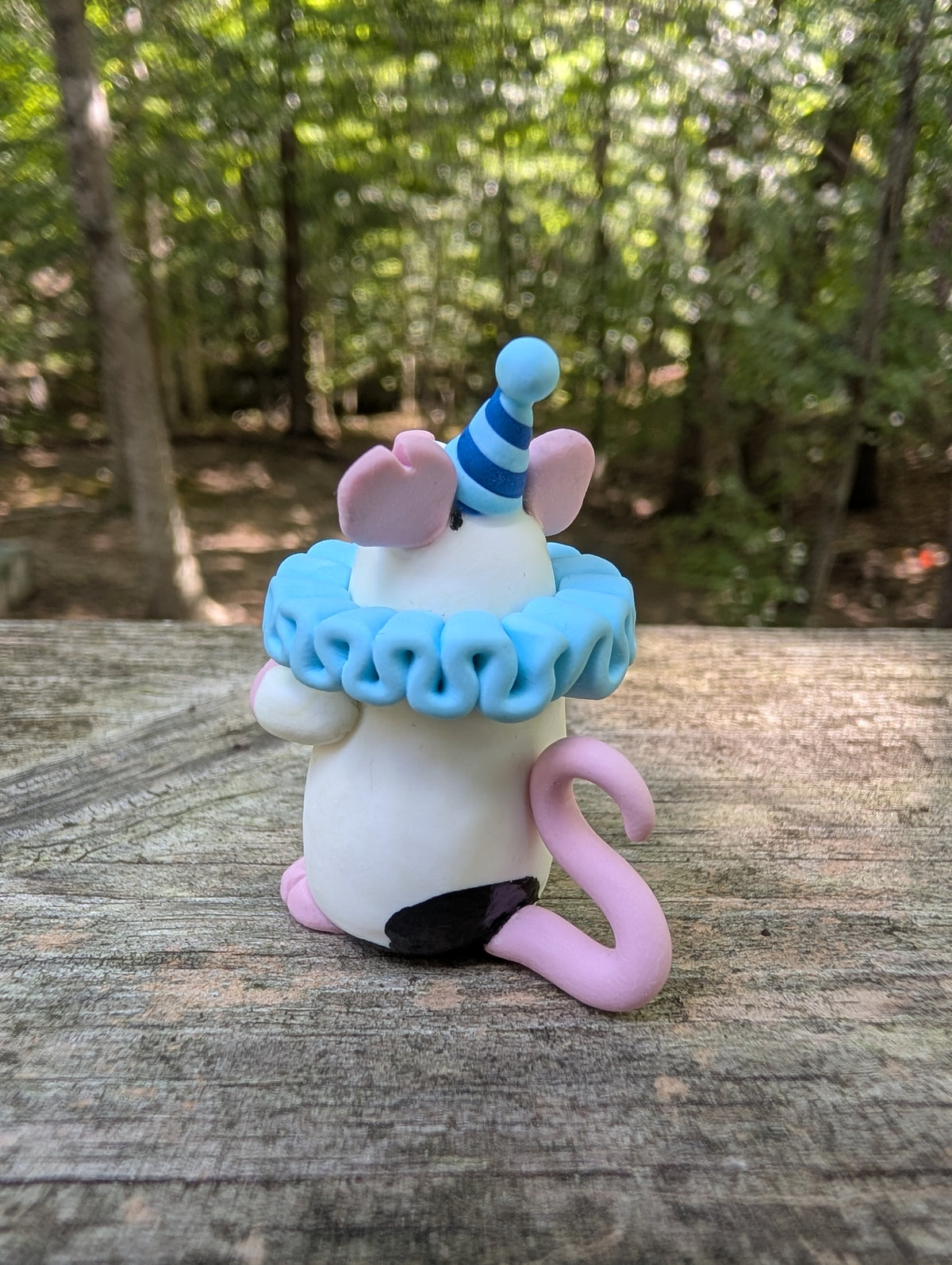 A small white mouse sculpture with pink ears and feet, black markings on the head and butt, wearing a blue striped party hat and a light blue ruffled collar. The mouse stands on it's back legs with it's hands in the air.