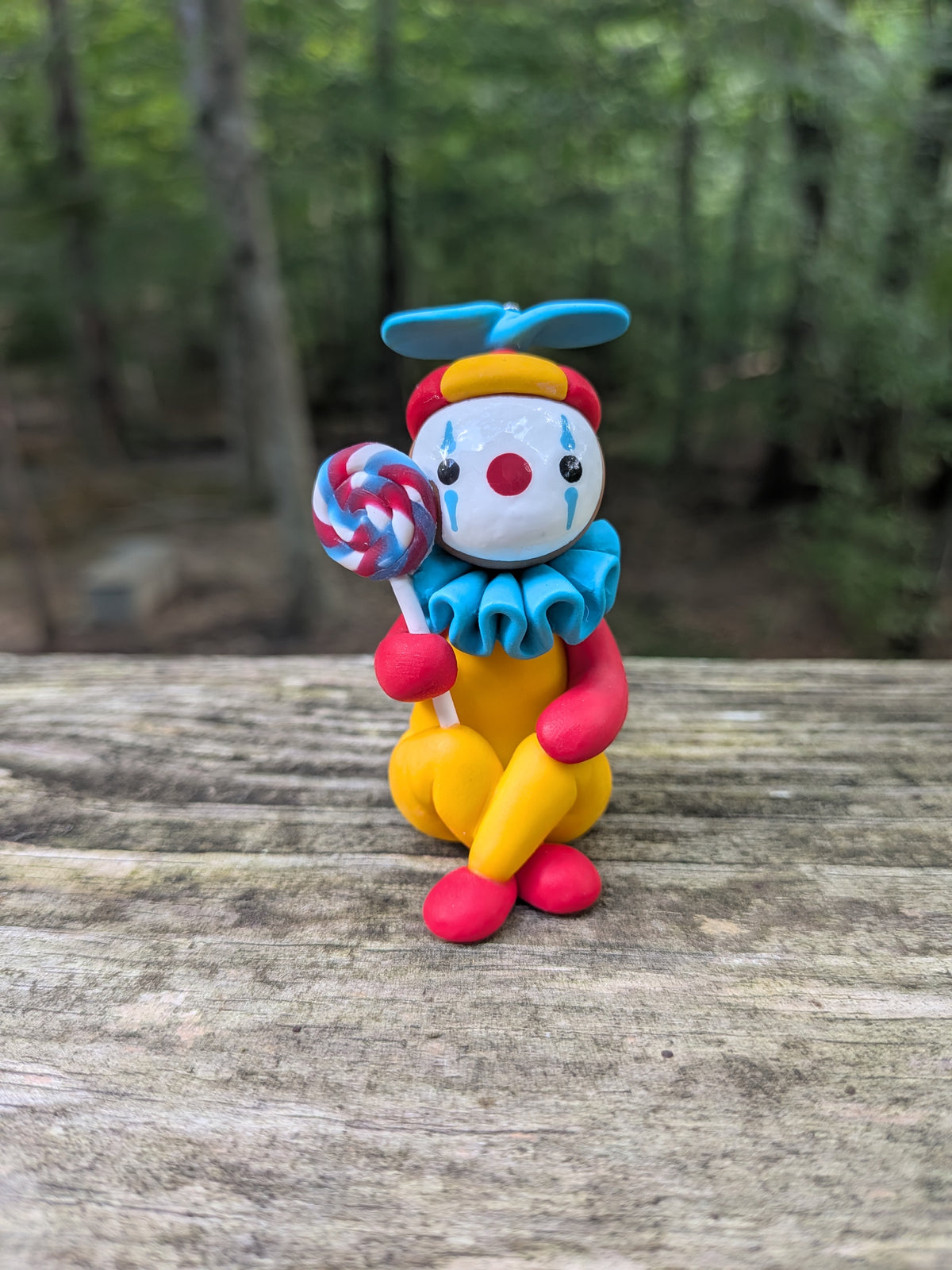 A clown sculpture. The clown yellow, red, and blue. They are holding a large swirly lolipop and sitting on the floor with their legs crossed.